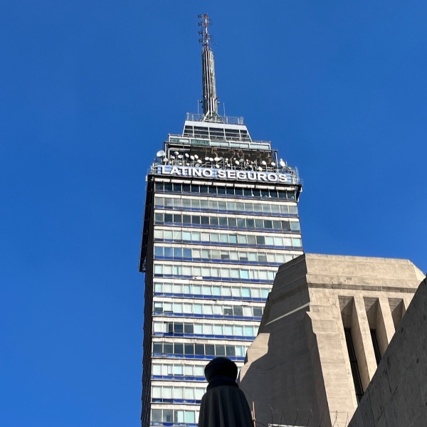 A low-angle view of the Torre Latinoamericana in Mexico City, with a clear blue sky in the background. The "Latino Seguros" sign is visible near the top, along with antennas and glass windows reflecting sunlight. A statue and part of another building are in the foreground.