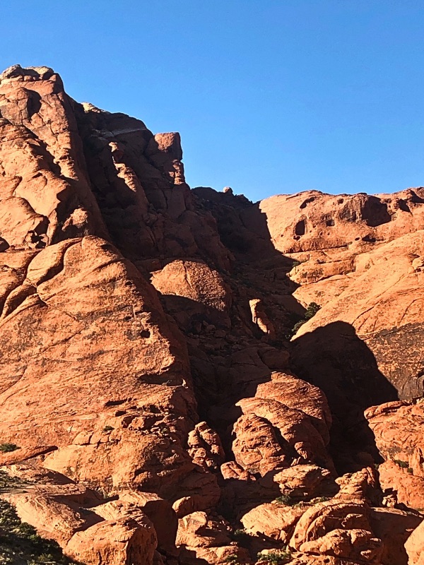 Rugged reddish-brown rock formations in the Mojave Desert under a clear blue sky, with deep crevices, jagged edges, and sunlight casting sharp shadows on the terrain.