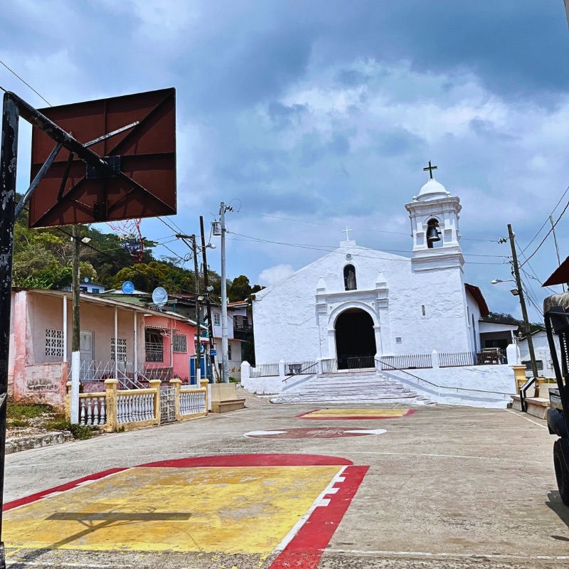 A white colonial-style church with a bell tower and cross stands at the center of a small town square on Taboga Island, Panama. In the foreground, a worn basketball court with red and yellow markings contrasts with the church. Surrounding the area are colorful houses, utility poles, and lush green hills under a cloudy sky.