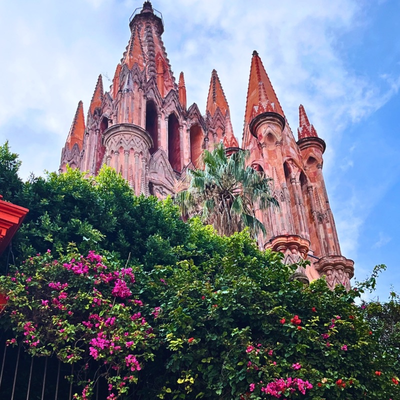 The iconic Parroquia de San Miguel Arcángel in San Miguel de Allende, Mexico, towering against a bright blue sky, surrounded by vibrant bougainvillea flowers.
