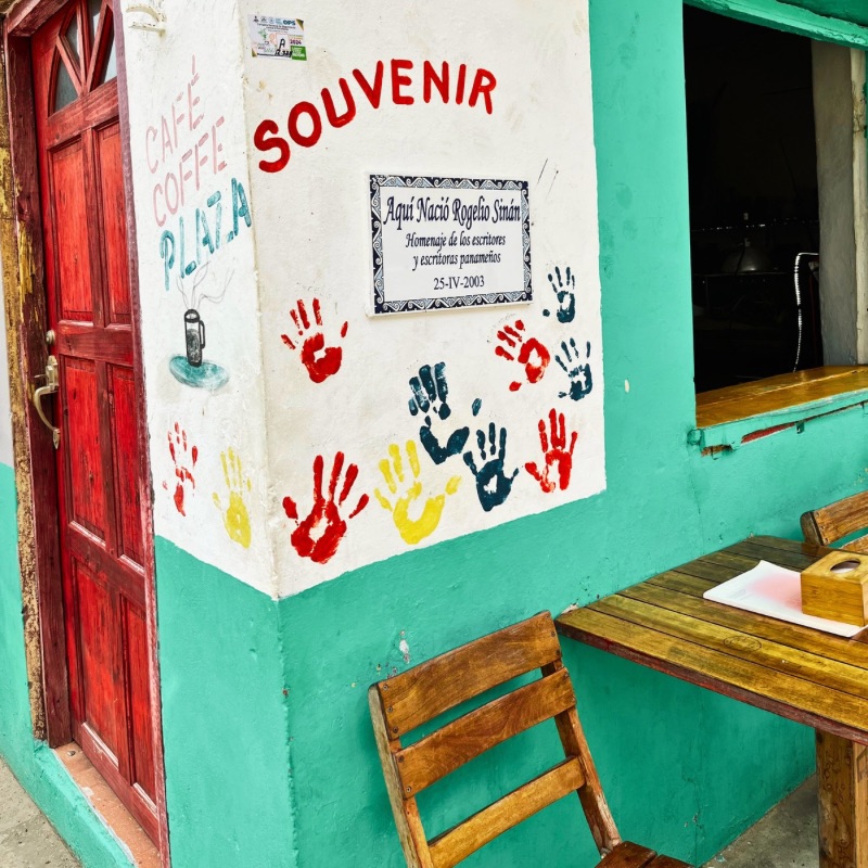 Partial front view of a coffee shop on Taboga Island, Panama, with a white and green wall decorated with colorful handprints. A plaque marks it as the birthplace of Rogelio Sinan, a celebrated Panamanian writer known for his modernist and surrealist literature.