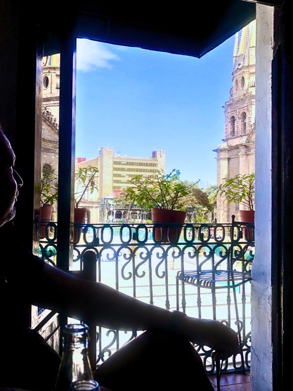 A silhouette of a person sitting near a balcony with a bottle in the foreground, overlooking the main plaza of downtown Guadalajara. The Guadalajara Cathedral with its iconic twin towers is visible in the background, along with potted plants on the railing and a clear blue sky.
