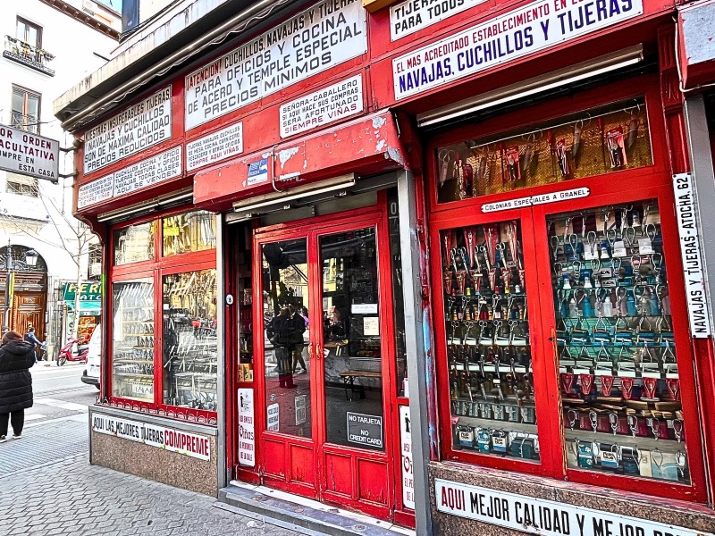Exterior image of Cuchilleria Viñas. The shop is filled with signs, predominantly in Spanish, advertising knives, scissors, and other cutting tools. The window displays showcase a variety of these tools, neatly arranged in rows. The entrance has a glass door, through which the interior of the store is partially visible. The shop is located on a busy street with pedestrians and surrounding buildings in the background.