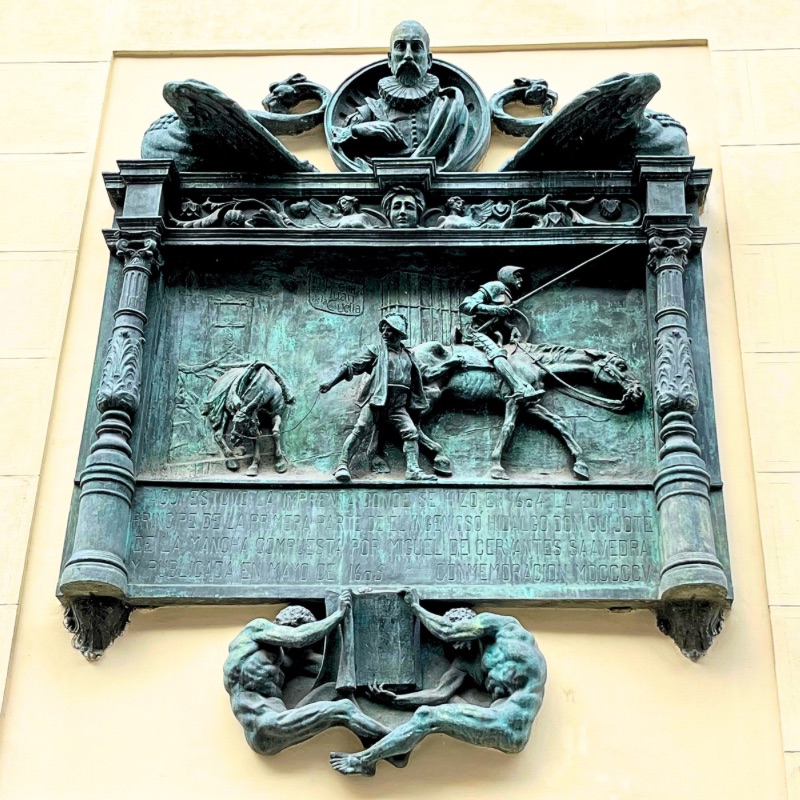 A commemorative plaque on a historic brick wall at Calle de Atocha 87, Madrid, marking the site where Don Quixote was first printed. The engraved words honor Miguel de Cervantes and the novel that changed literature forever.