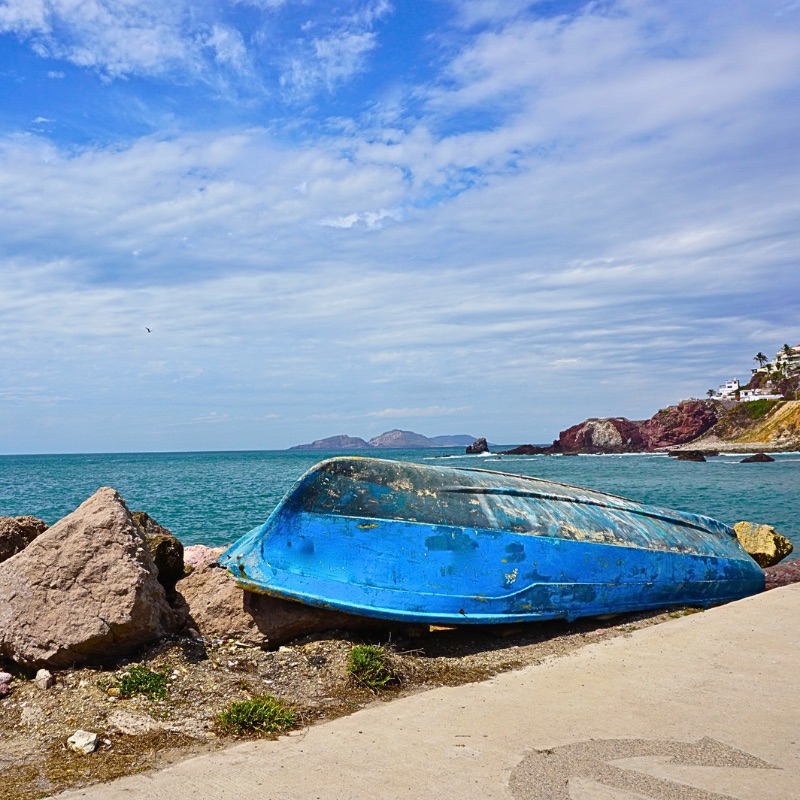 A weathered blue boat overturned on the shore, its worn wood facing the sky. In the distance, Mazatlán’s coastline curves along the sea, while the ocean glows in deep hues of turquoise.