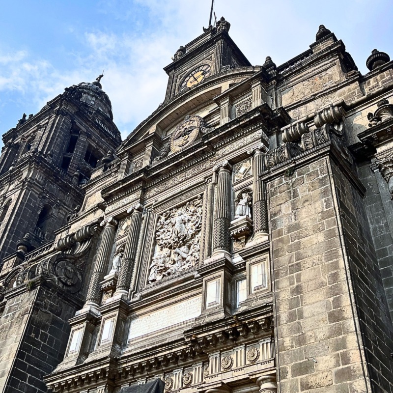 Low-angle view of the Metropolitan Cathedral in Mexico City, showcasing its intricate stone façade and sculpted figures against a vibrant blue sky.
