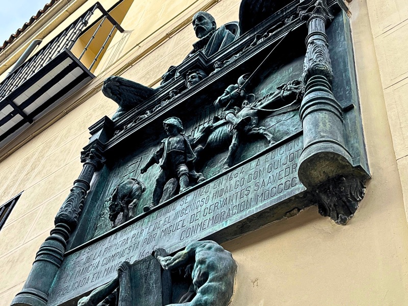 A bronze relief sculpture mounted on a beige wall depicts Miguel de Cervantes at the top, overlooking a scene from Don Quixote. Below, Don Quixote strides forward in armor, with Sancho Panza following behind. In the background, windmills appear as looming figures. The sculpture includes an inscription commemorating the first edition of Don Quixote, marking the location where Don Quixote was born in print.