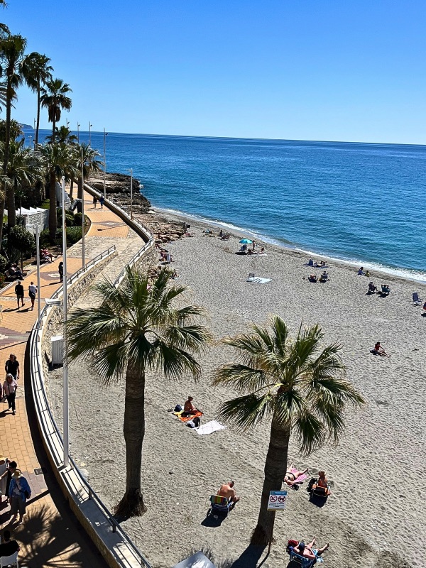 The deep blue sea glistens under the sun at La Torrecilla beach in Nerja. A few visitors walk along the shoreline, their footprints fading into the dark, soft sand.