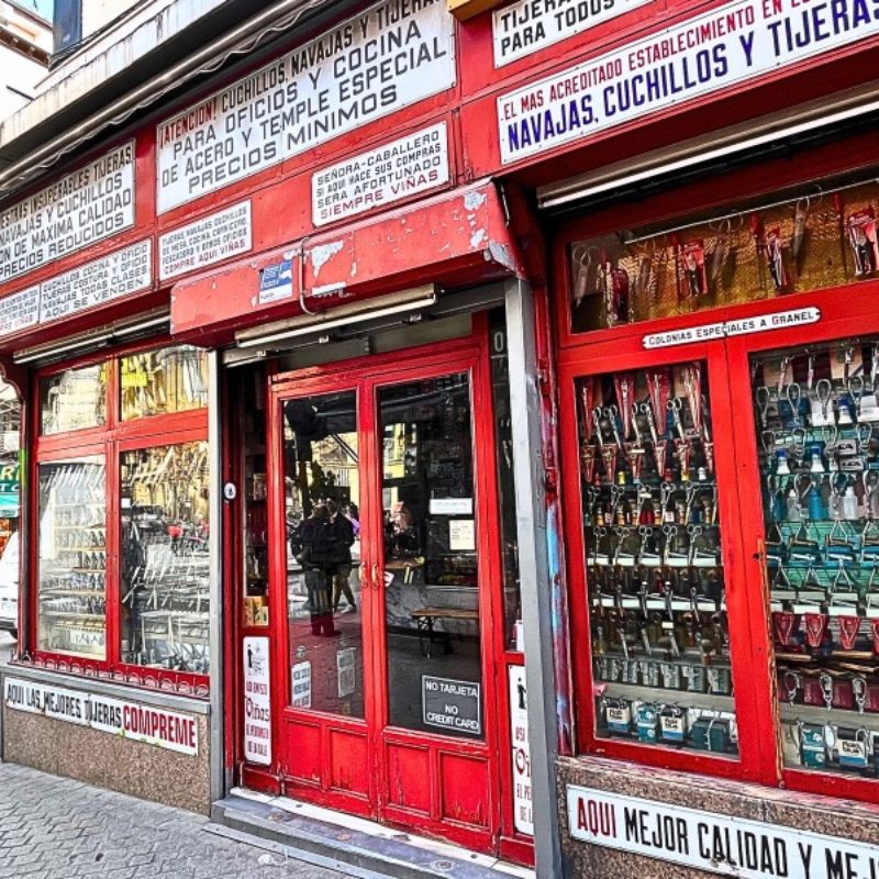 A historic cutlery shop, Cuchillería Viñas, at Calle de Atocha 62 in Madrid, with a red storefront, vintage signage, and window displays filled with knives and scissors. The shop's worn exterior reflects its century-old history, while pedestrians walk past on a lively Madrid street.
