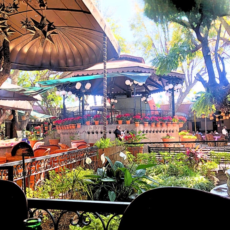 A vibrant outdoor restaurant in Tlaquepaque’s main square, surrounded by lush greenery, terracotta pots filled with flowers, and ornate wrought iron railings.