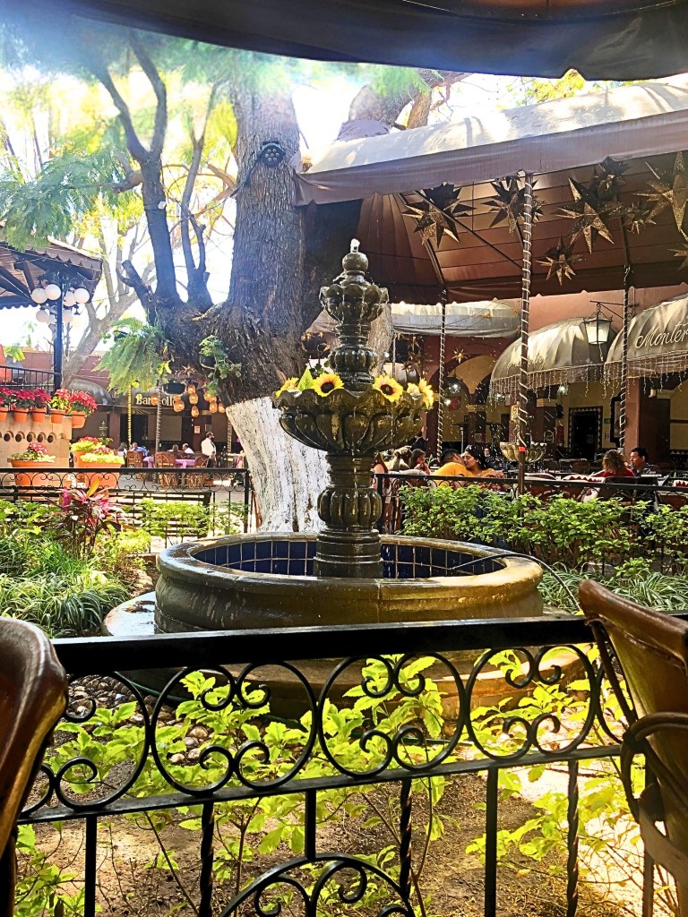 A sunlit courtyard in Tlaquepaque’s main square featuring a tiered stone fountain adorned with sunflowers, surrounded by greenery and wrought-iron railings.