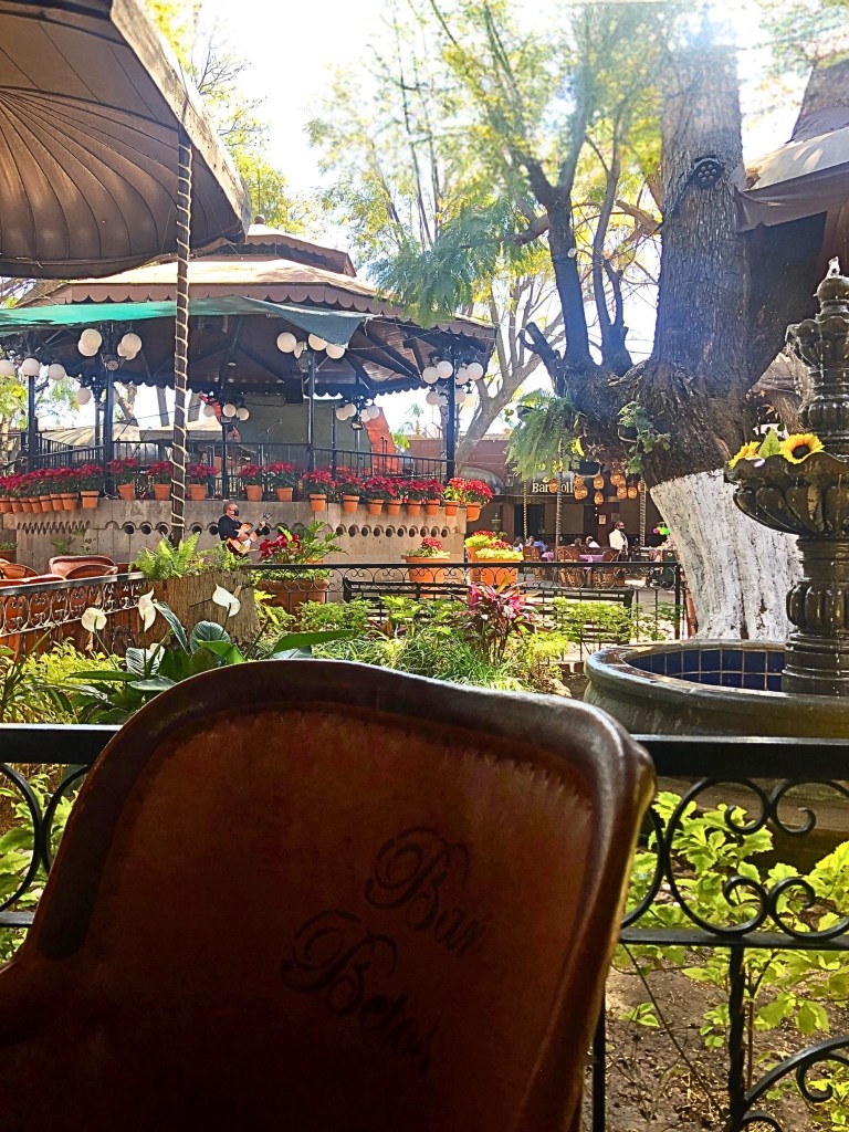 Sunlit patio with leather “equípales” chairs, hanging lanterns, and scalloped canopies providing shade, set in a picturesque garden with blooming plants.  
