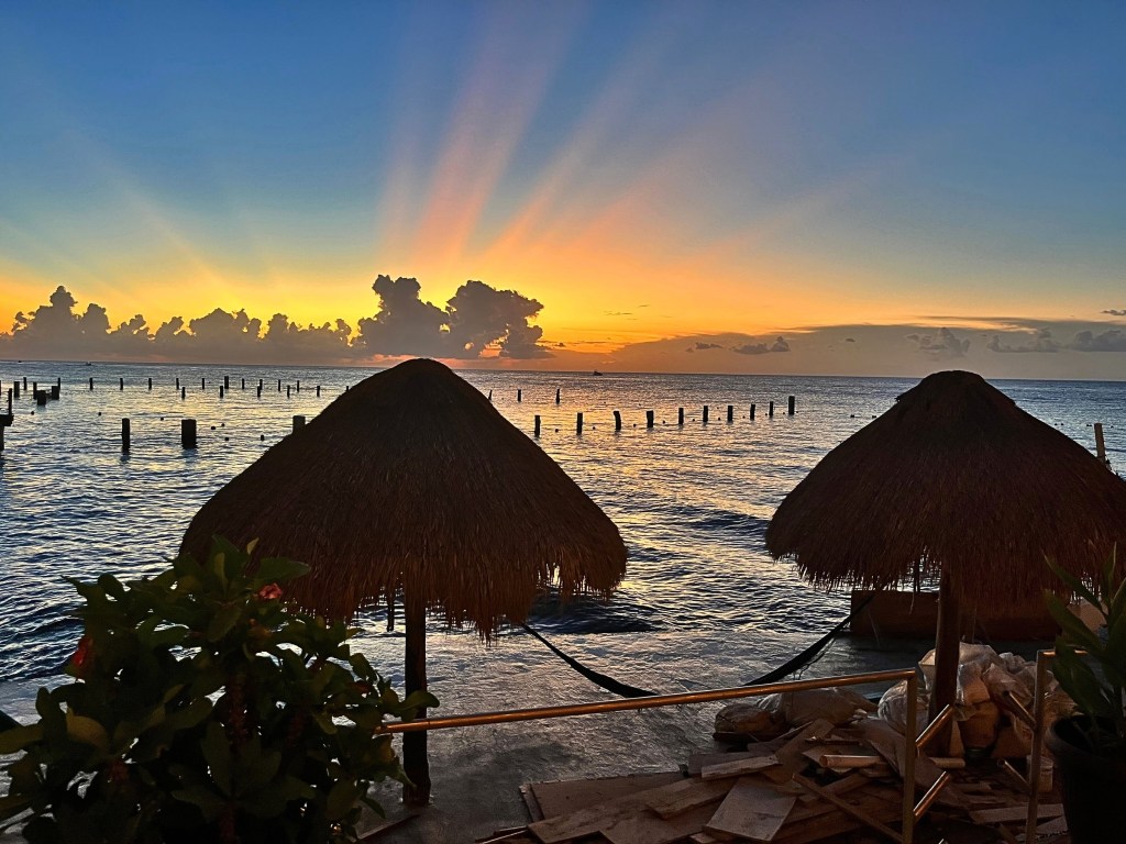 Two hammocks beneath thatched roofs at the edge of the sea during a glowing sunset in Cozumel.