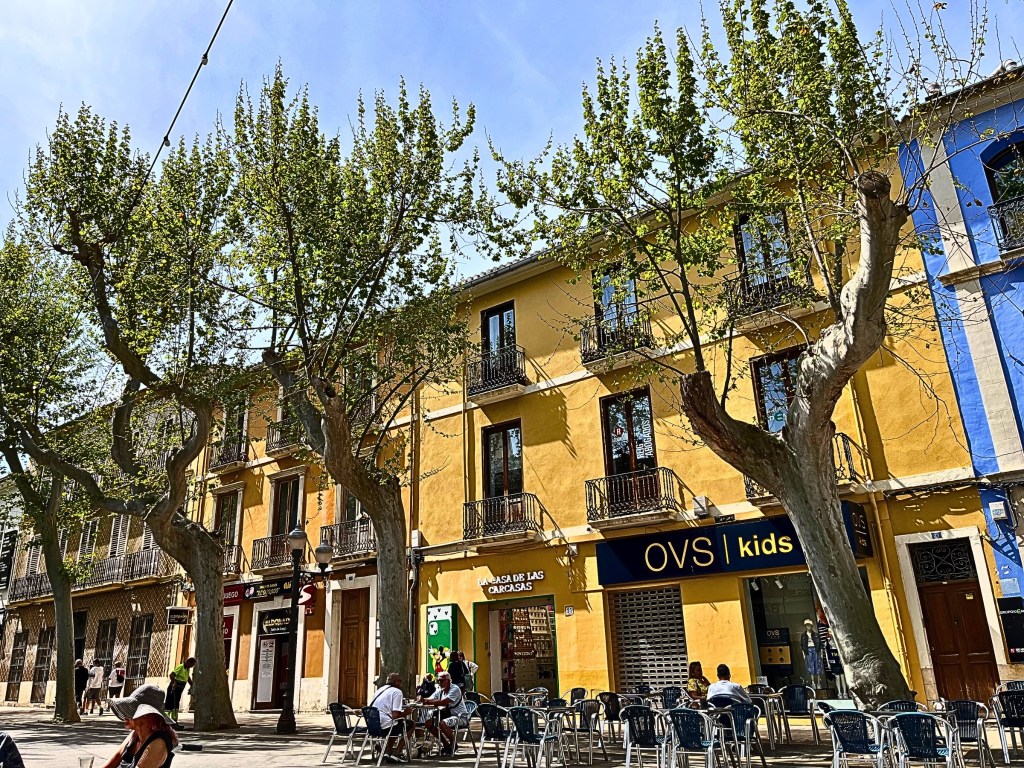 Golden-hued building façade in Dénia, Spain, with ornate wrought-iron balconies and two white doves perched peacefully in the afternoon sun.