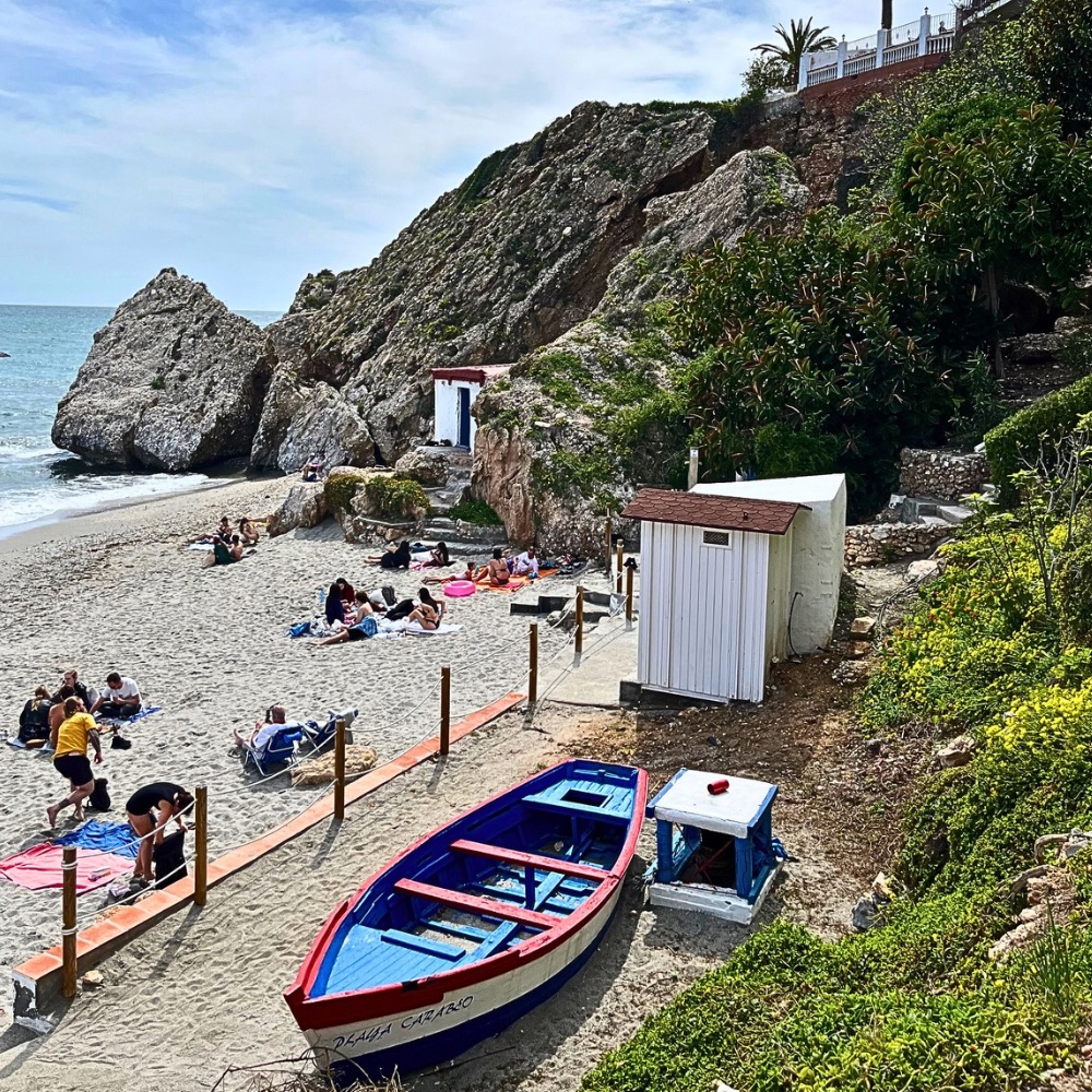 View of Playa Carabeo beach in Nerja, Spain from a stair-access path, with rocks, sand, and the sea in the background.