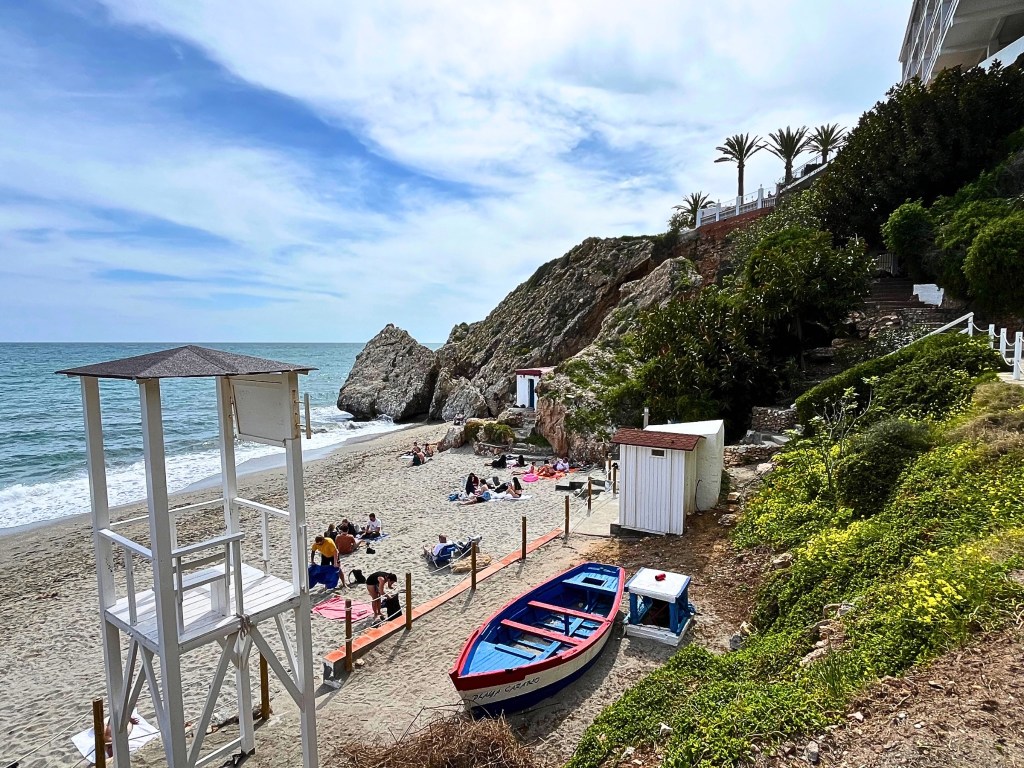 A lifeguard tower overlooks a beach dotted with people and bordered by rocky cliffs and blue ocean.
