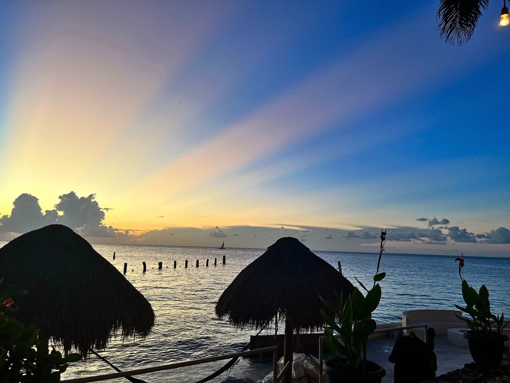 Sunset over Cozumel with light rays fanning from clouds and palapa huts in the foreground.