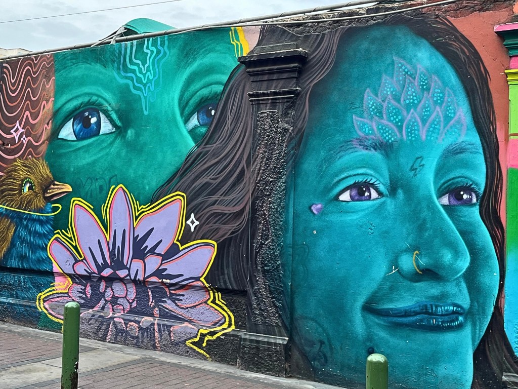 Close-up of an urban wall painting showing ethereal human faces, a bird, and a glowing lotus in bold, dreamy hues. 