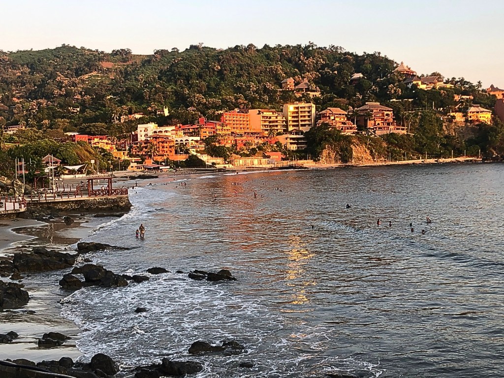 View of Zihuatanejo bay at sunset with sunlit buildings and swimmers in the calm sea.