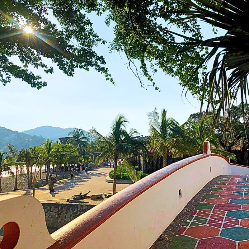 A colorful tiled pedestrian bridge under tropical trees in Paseo del Pescador, Zihuatanejo, bathed in soft sunlight.