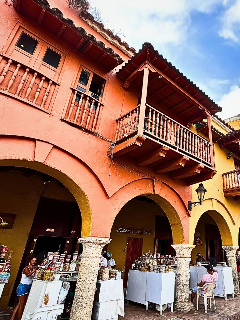 Close-up of a terracotta-orange building in Cartagena’s Walled City with street vendors selling candies and souvenirs under a shaded sidewalk.