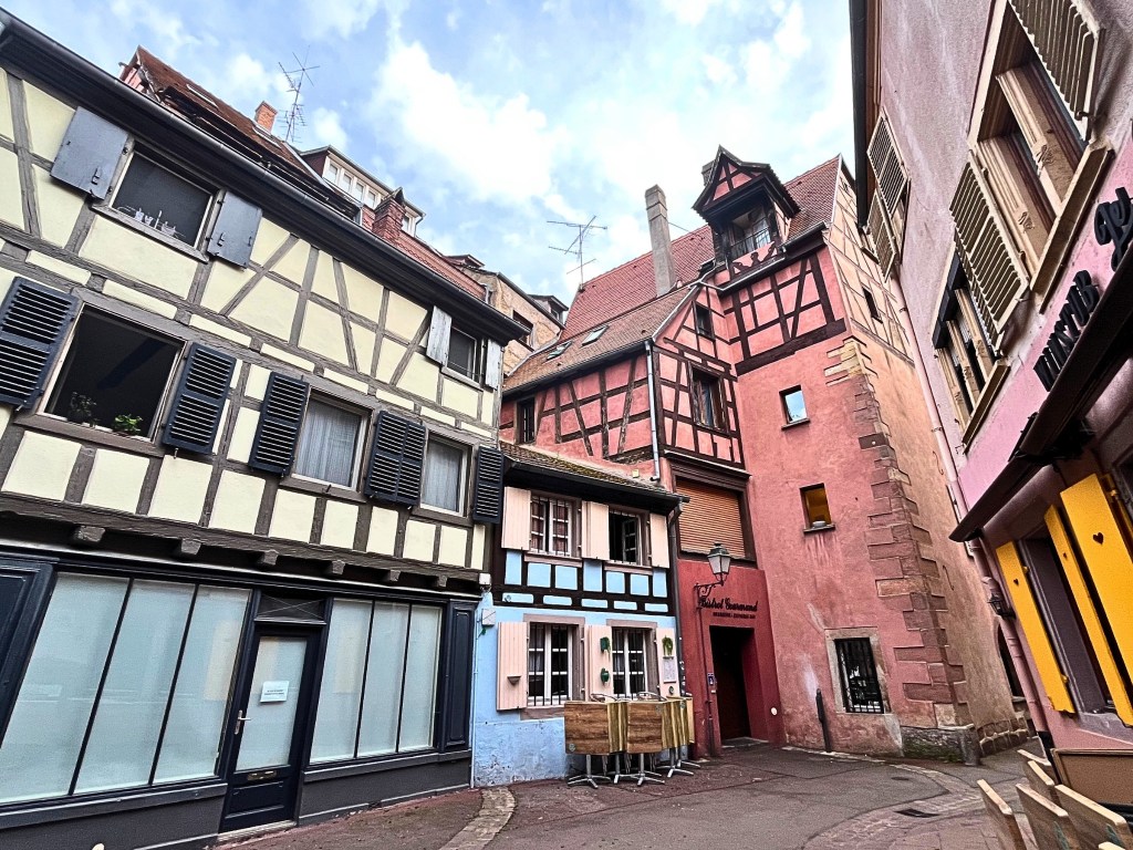 Narrow street in Colmar’s old town lined with pastel half-timbered buildings, including a blue house with shutters, a pink building labeled “Le Palais Gourmand,” and cozy café seating on cobblestones.
