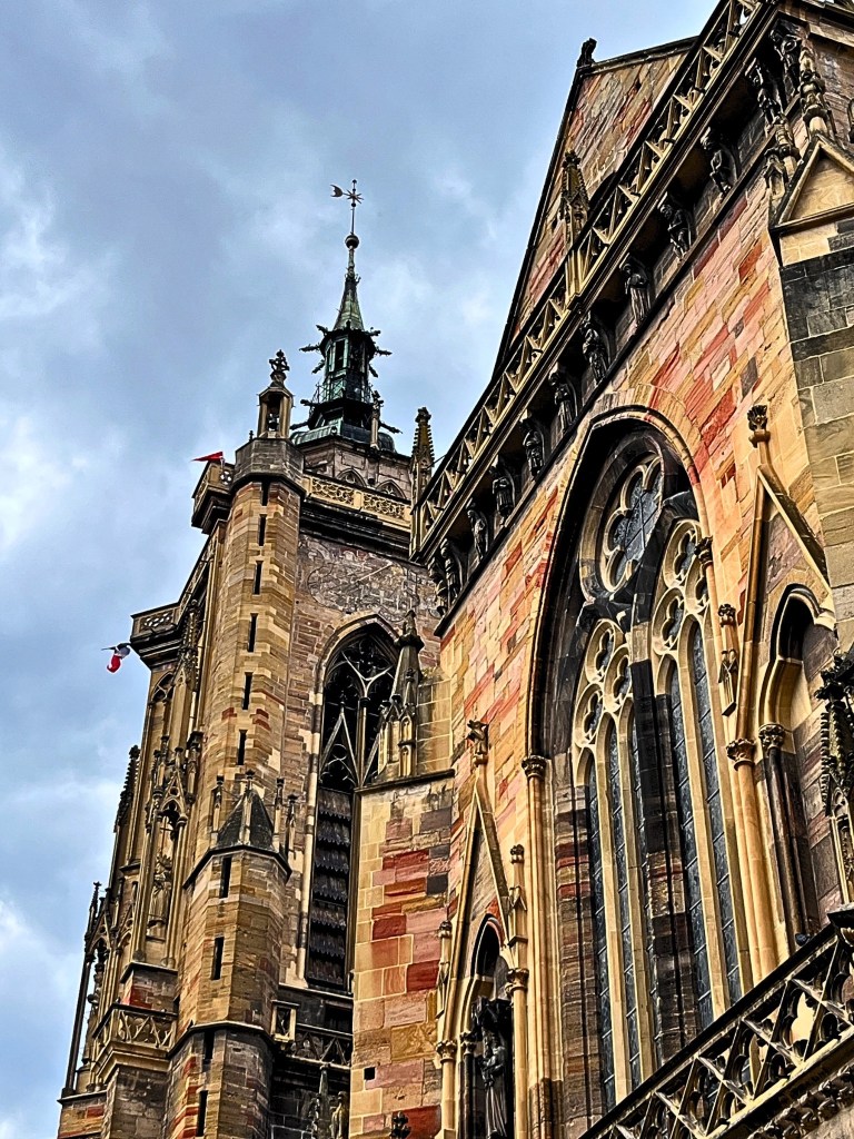 Side-angled view from the ground of the Collégiale Saint-Martin’s bell tower and lateral church wall, showing stained-glass windows, pointed arches, and sculpted stone details.