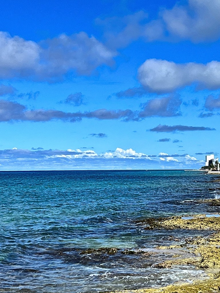 Cozumel shoreline with open sea on the left, showing a gradient of water colors from clear to jade, turquoise, and deep blue; limestone shore on the right, with a building in the distance and blue sky with scattered clouds above.