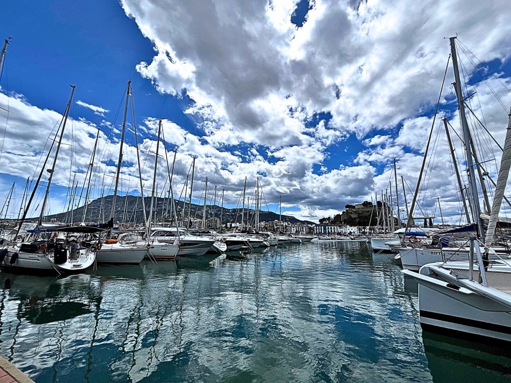 A calm marina in Dénia with over a dozen yachts at rest; their masts reflected in still water beneath a blue sky with drifting clouds. In the background, Dénia Castle stands on a hill to the right.