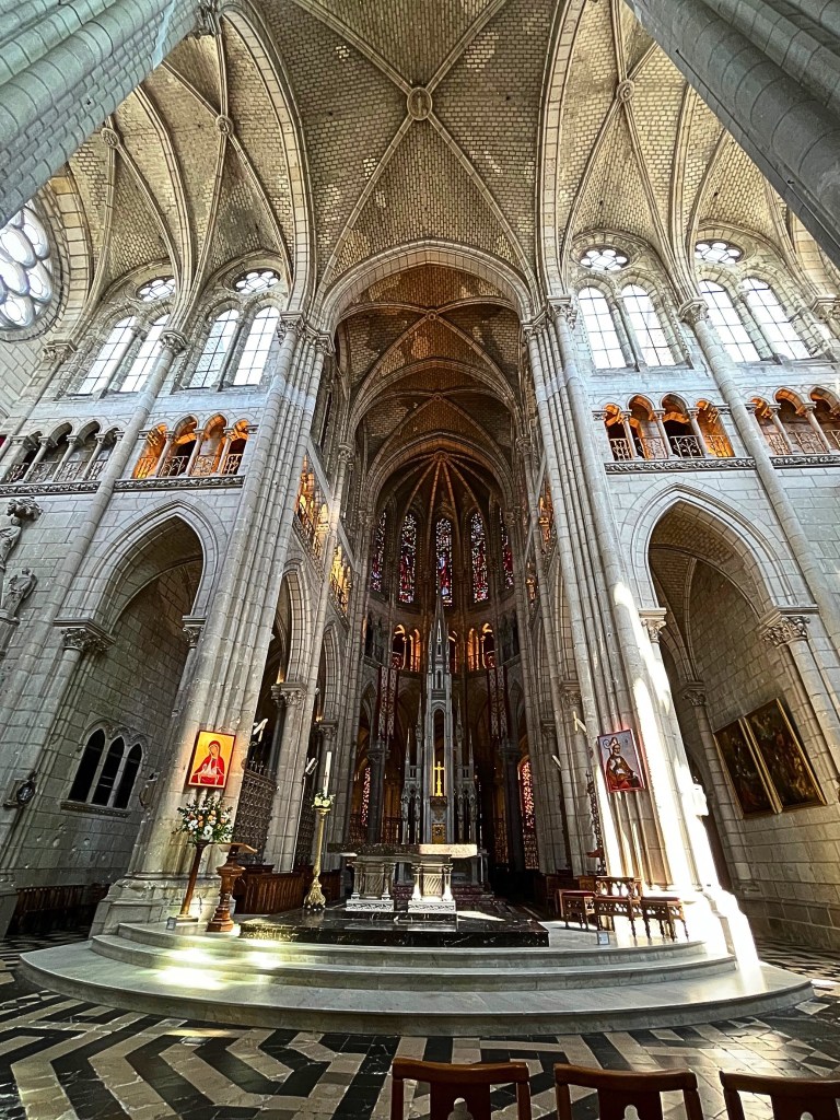 Central nave of the Saint-Nicolas Basilica in Nantes with vaulted Gothic ceilings, religious iconography in stained glass, and an illuminated high altar.