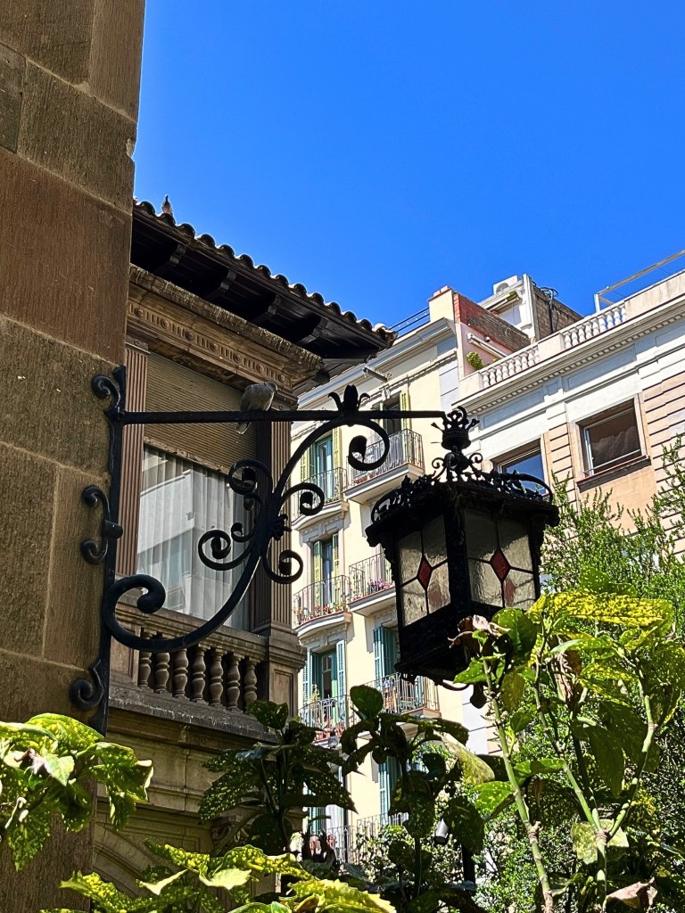 Wrought-iron lantern with stained glass panels mounted on a stone villa wall in Jardins de Muñoz Ramonet, Barcelona, with lush greenery and pastel apartment buildings in the background under a clear blue sky.