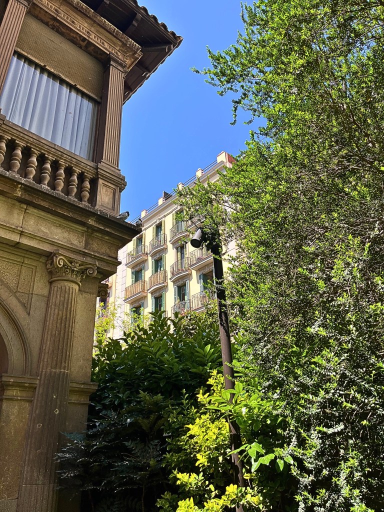Warm-toned stone façade of an early 20th-century villa in Jardins de Muñoz Ramonet, Barcelona, with carved details, wooden eaves, and lush greenery in the foreground; pastel apartment buildings and a blue sky in the background.