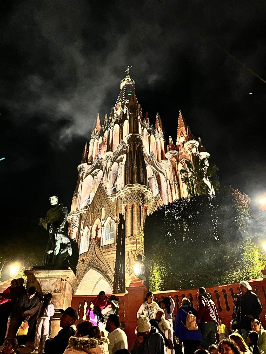 The Parroquia de San Miguel Arcángel glows against a smoky, dark sky during La Alborada in San Miguel de Allende. A crowd stands before the church, wrapped in warm clothing, while a bronze statue of a monk watches over the scene. The church's spires reach upward, framed by smoke from the fireworks that have just erupted.