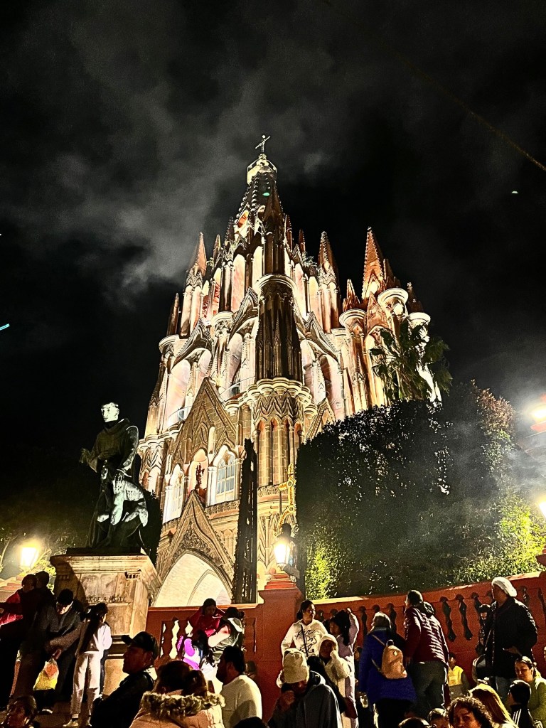The Parroquia de San Miguel Arcángel glows against a smoky, dark sky during La Alborada in San Miguel de Allende. A crowd stands before the church, wrapped in warm clothing, while a bronze statue of a monk watches over the scene. The church's spires reach upward, framed by smoke from the fireworks that have just erupted.