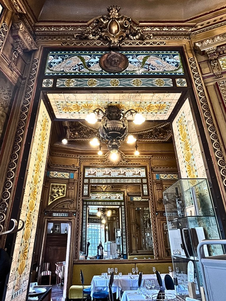 A grand chandelier hanging from a painted ceiling, casting a soft glow over the brasserie’s richly decorated dining room.