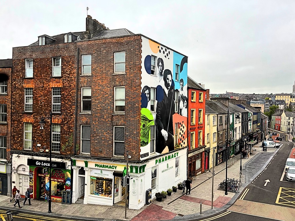 Corner brick building at the intersection of MacCurtain Street and St. Patrick’s Hill in Cork, Ireland, featuring a ground-floor pharmacy, tall red-brick facade, and a large mural of expressive painted faces covering the upper side wall — a striking urban art landmark in the heart of the city.
