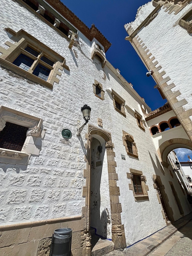 Angled ground-level view of the right side of Palau Maricel in Sitges, showing a small arched passageway, simple elegant windows along a white brick wall, and part of the tower on the far right, all beneath a vivid blue sky.