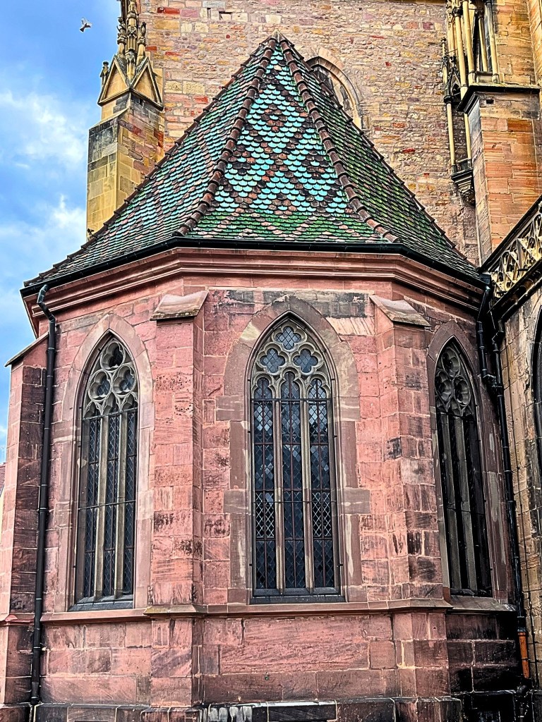 Close-up of the rear tower of the Collégiale Saint-Martin, showing soft rose stone and a roof with green, blue, and muted gold geometric tile patterns.