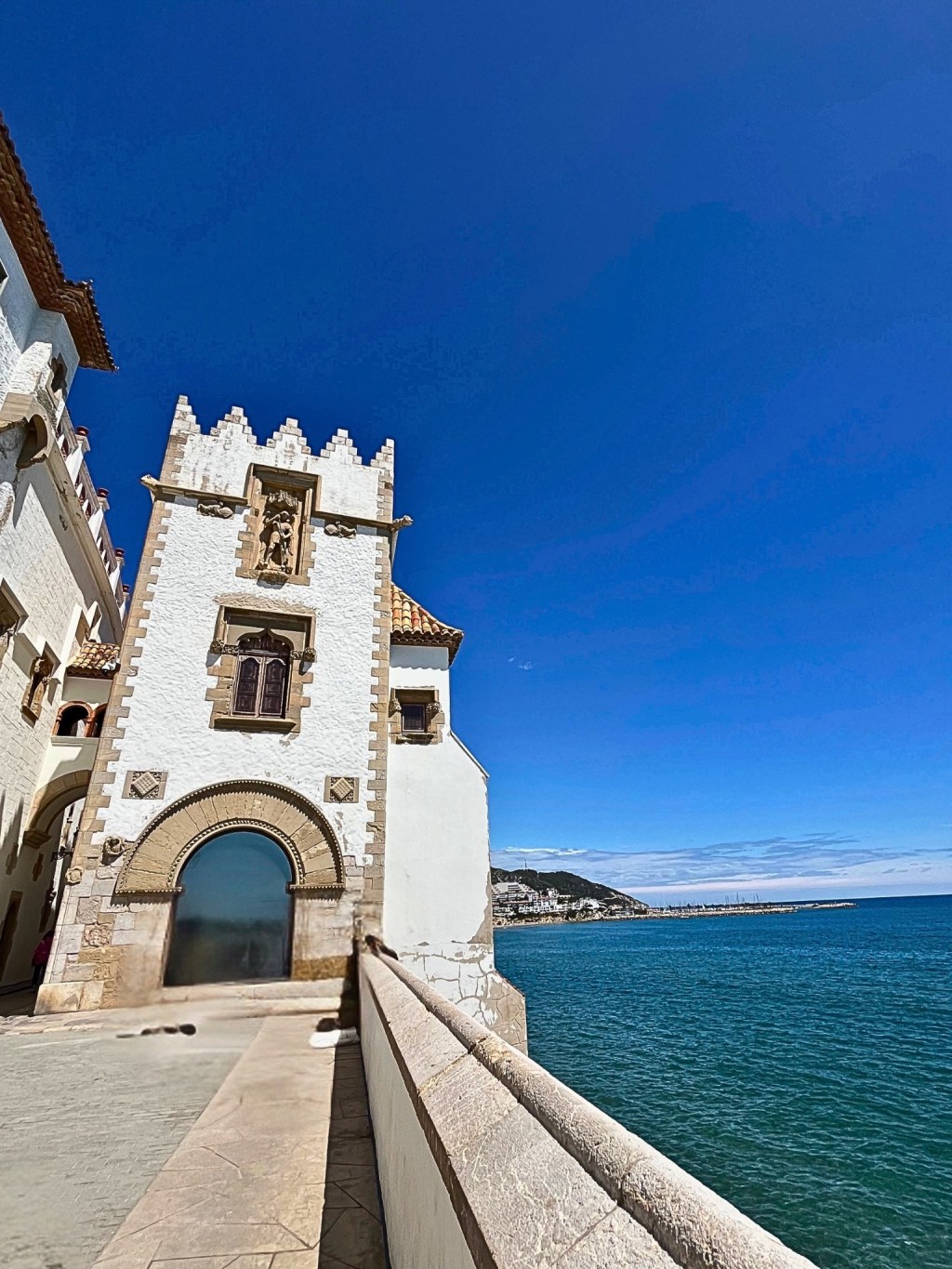The white tower of Palau Maricel stands in quiet grandeur, facing the Mediterranean on its right. Built from blocks of natural stone at the base, the tower ascends through a graceful semicircular arch framed with delicate carvings, a single Moorish-inspired window, and a sculpted crown embedded in stone. At its top, a geometric frieze of white brick triangles reaches toward the deep blue sky. The sea glimmers to the right, echoing the calm strength of this seaside structure.