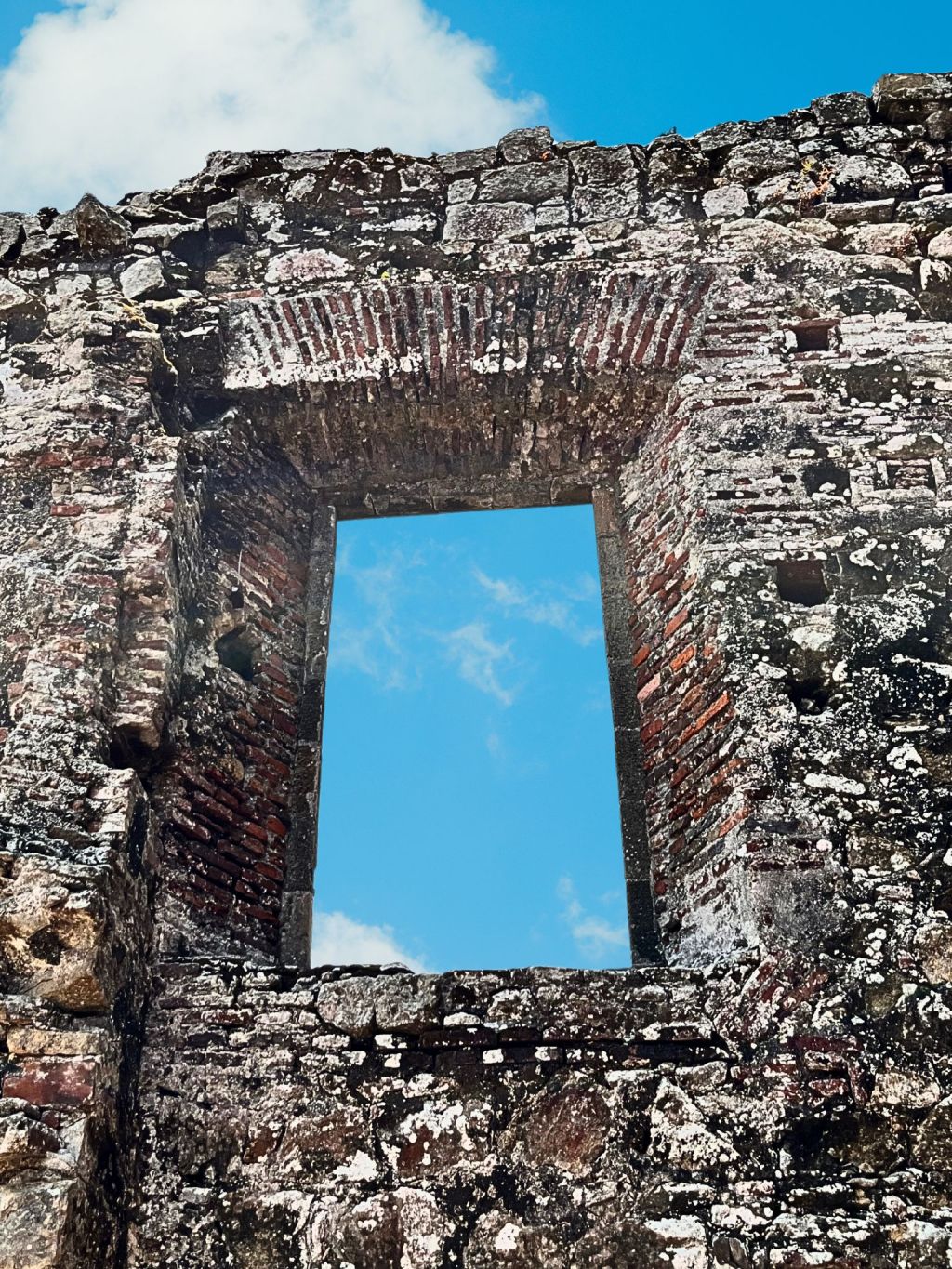 Close-up of a ruined window in Old Panama City, showing coquina, bricks, and mortar with patches of white lichen and a view of blue sky through the opening.