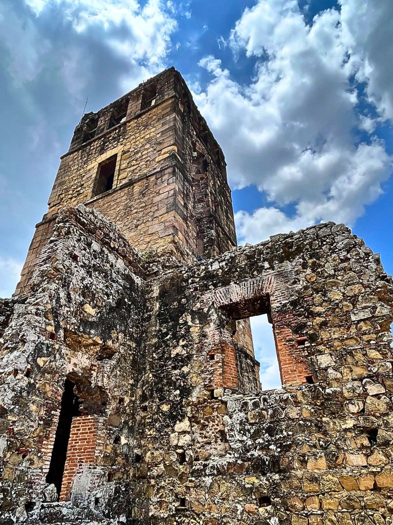Stone bell tower of the Cathedral of Nuestra Señora de la Asunción in Panamá Viejo, standing tall against a bright blue sky with scattered white clouds.