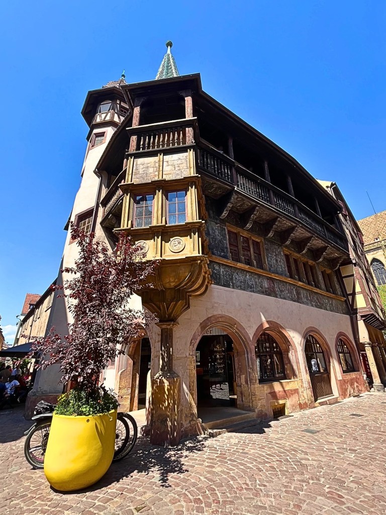 A dramatic upward shot of the Pfister House in Colmar, France, highlighting its Renaissance architecture with a turret, oriel window, timber balcony, and a vivid yellow planter in the foreground.