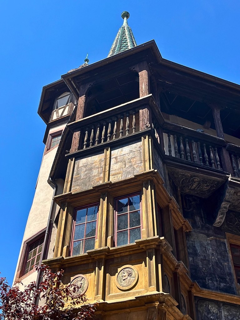 Upward-angled photo of the Pfister House in Colmar, France, showing a honey-colored oriel window, faded Renaissance murals, a wooden loggia, and a green-tiled turret under a clear blue sky.