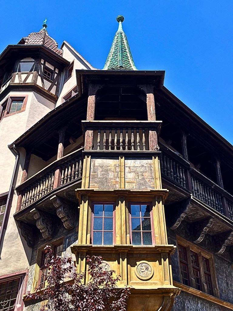 A close-up view of the Pfister House in Colmar, focusing on its golden ochre polygonal oriel window, ornate wooden panels, carved medallions, faded mural paintings, wooden loggia, and turreted roofs, all against a cobalt blue sky.