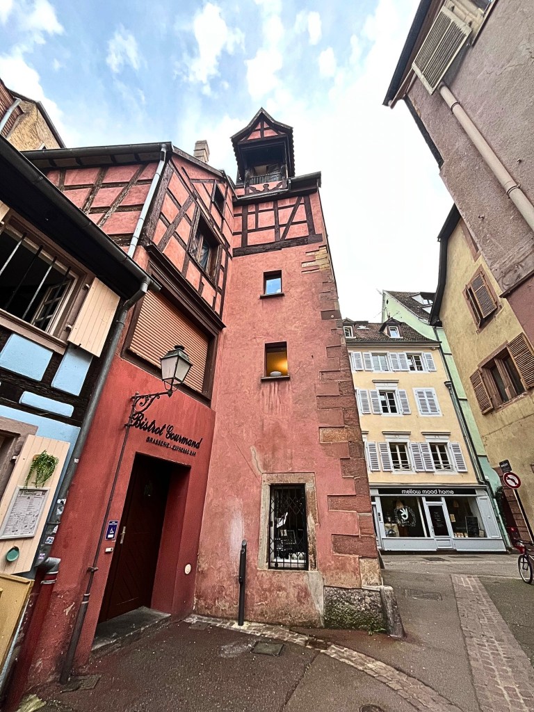 Close-up ground-level view of Le Palais Gourmand in Colmar, showing pink half-timbered facade with a steep roof and ornate gabled window, framed by nearby yellow and mustard buildings with white and brown shutters.