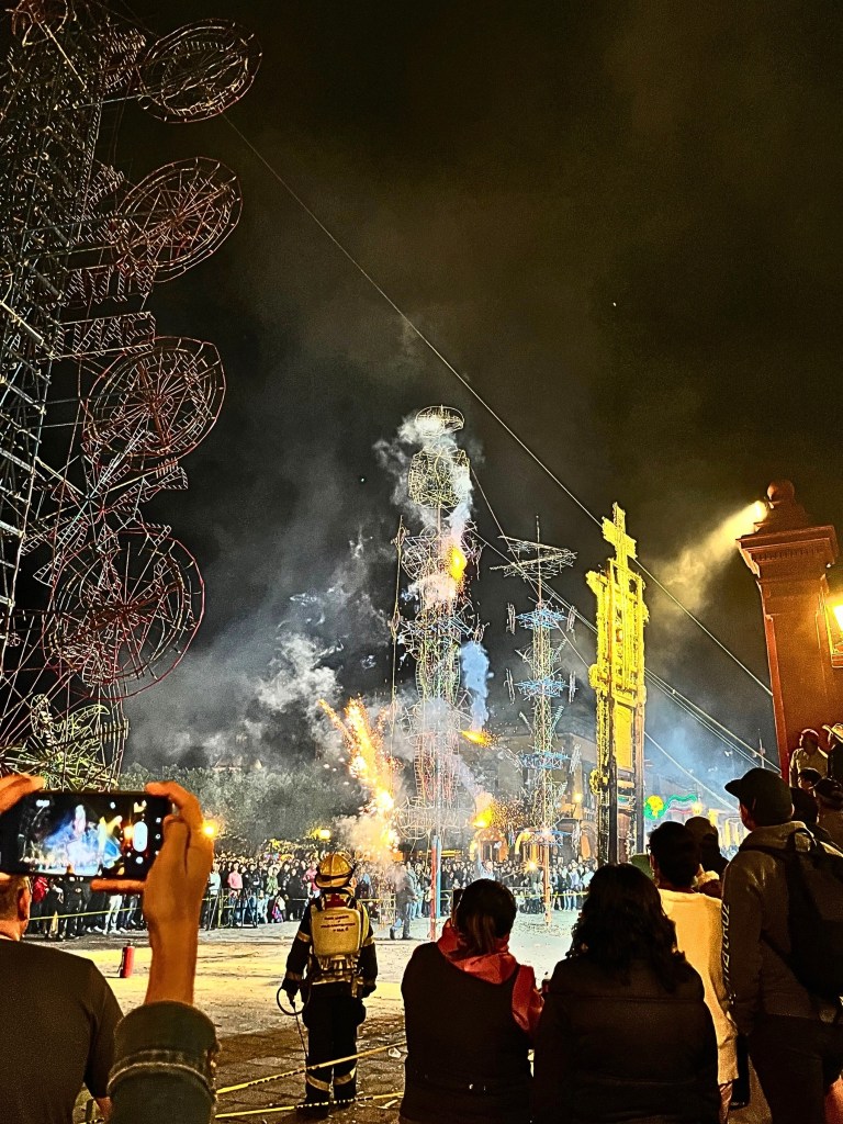  A crowd gathers in anticipation as tall, handcrafted firework towers (toritos) stand against the night sky, their colorful geometric patterns glowing softly. Sparks flicker from their base as smoke rises, with the silhouette of San Michael the Archangel traced in pyrotechnic wire. A firefighter stands in the foreground, ready for the eruption of fireworks during San Miguel de Allende's La Alborada celebration.