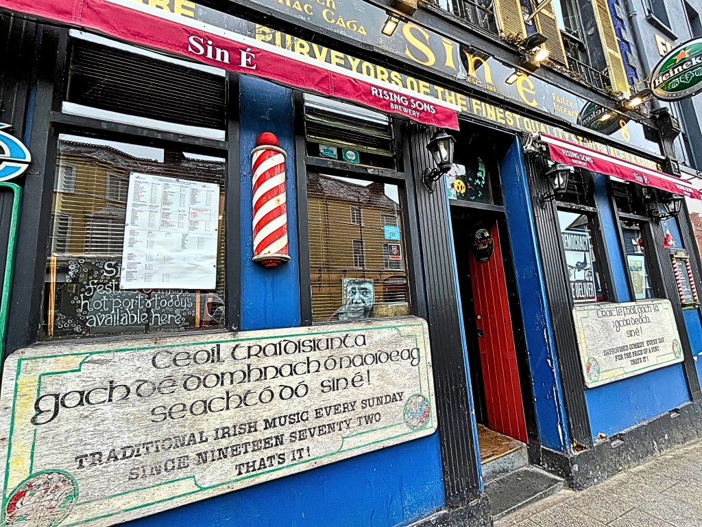 Photo of the colorful Sin É pub in Cork City, Ireland, with a deep blue façade, burgundy awning, and signage celebrating its traditional Irish music sessions since 1972.