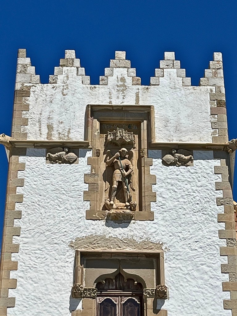 Close-up of the upper section of Palau Maricel’s white tower in Sitges, featuring a weathered stone sculpture of Saint Michael embedded near the top, and crowned with a distinctive zigzag frieze of white brick triangles that stand out crisply against the vivid blue Mediterranean sky.
