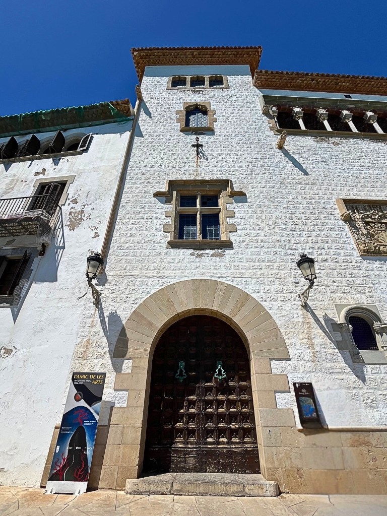 Main entrance of Palau Maricel museum in Sitges, with a natural stone arch framing an old carved wooden door with green iron knobs shaped like bells, flanked by lanterns; above, the white brick façade features layered windows and a terracotta-tiled roof, all set beneath a vivid blue sky.