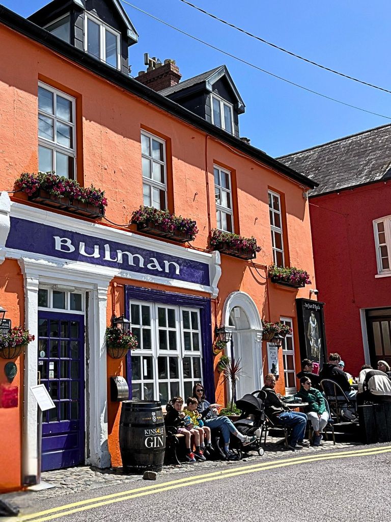 Bright orange exterior of The Bulman pub in Kinsale, with white-trimmed windows, a black door, and a purple sign with white lettering.